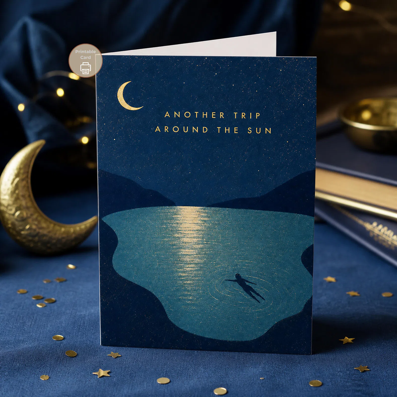 Styled product mockup: A printable birthday card with a midnight lake scene, golden crescent moon, starry sky, mountain silhouettes, and the message Another Trip Around The Sun.